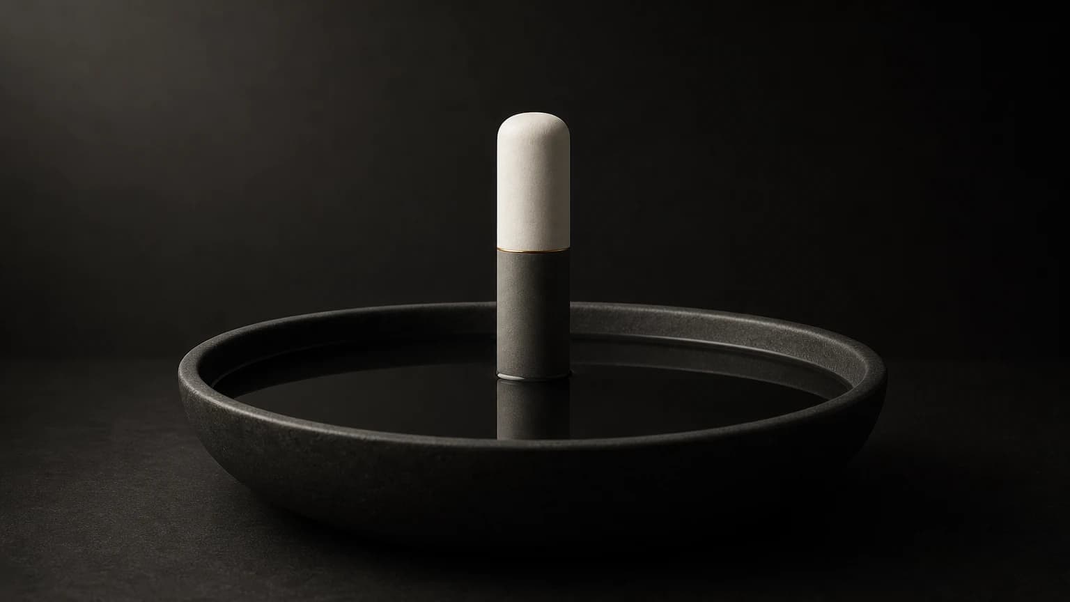 A slender matte column, ivory at its rounded top and dark gray below a thin brass ring, stands in the center of a shallow charcoal basin of still, dark liquid.